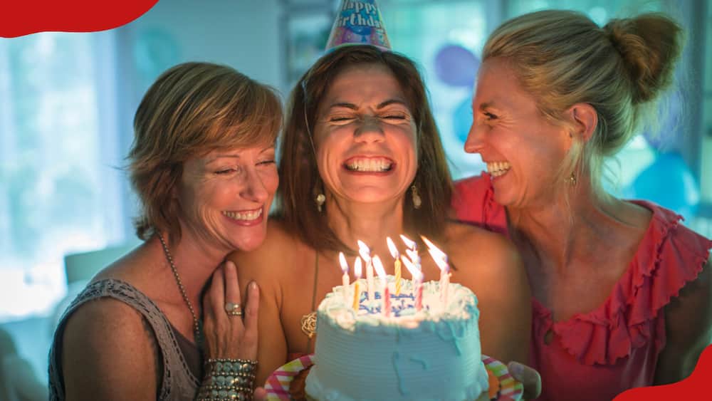 A mature woman holds a birthday cake and makes a wish while two women look on