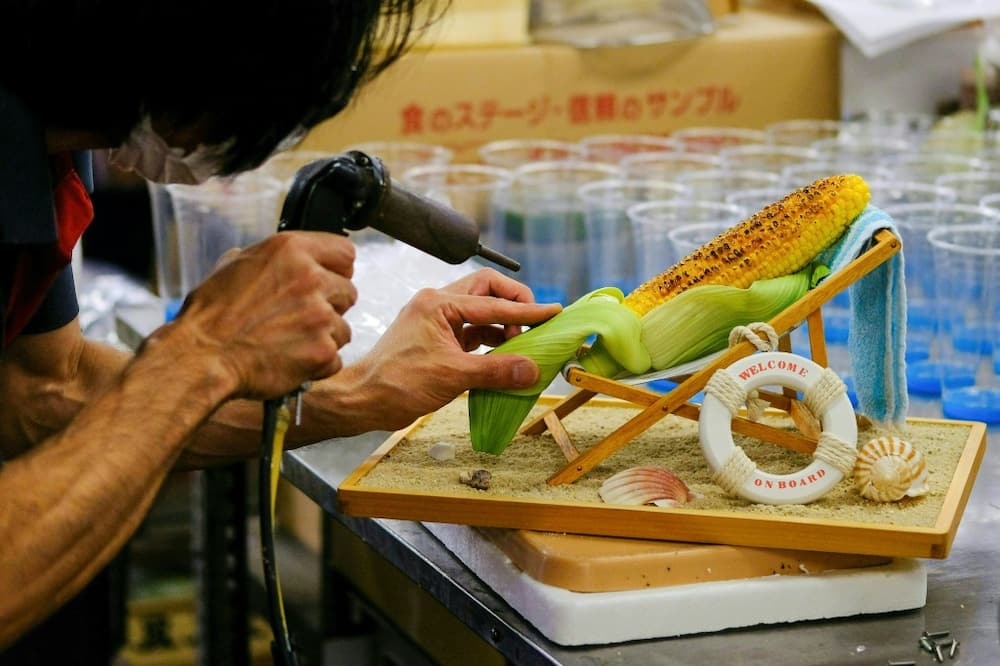 A worker applies the finishing touches to a plastic ear of grilled corn reclining on a beach chair