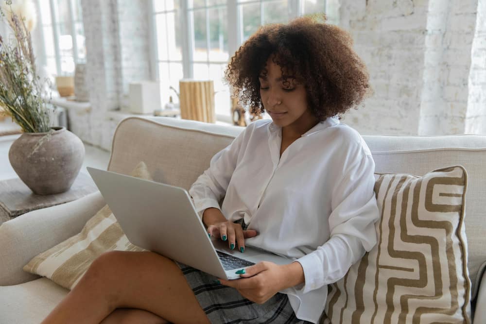 A young woman in a white shirt is using a laptop while sitting on a cosy sofa