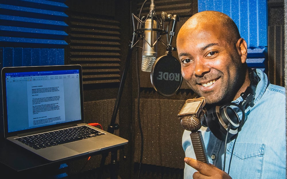  A bald black man smiles while sitting in front of a microphone and laptop with a document with the title 'Premier League Tactics: Formations, Pressing, and Counter-Pressing' on the screen.