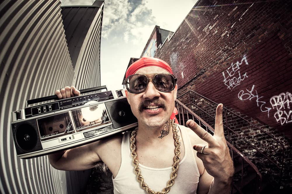 A hip hop / gangsta / rapper character with gold chains (bling), and doo rag stands in a graffiti covered alley, holding a large stereo cassette player (boom box) on his shoulder.