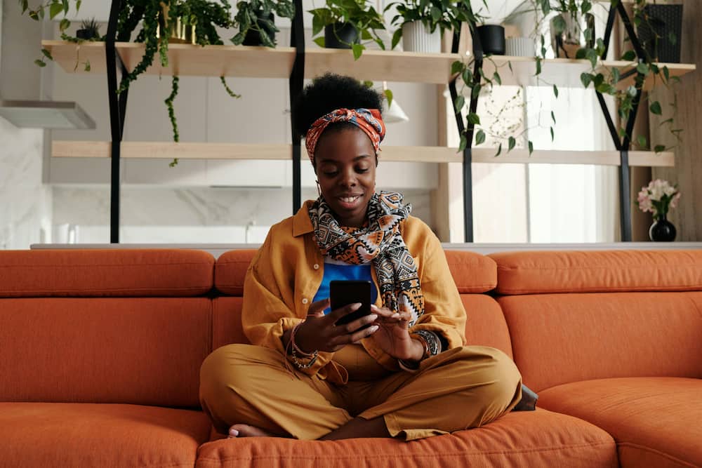 A black woman sitting on a couch smiling as she gazes on her phone.