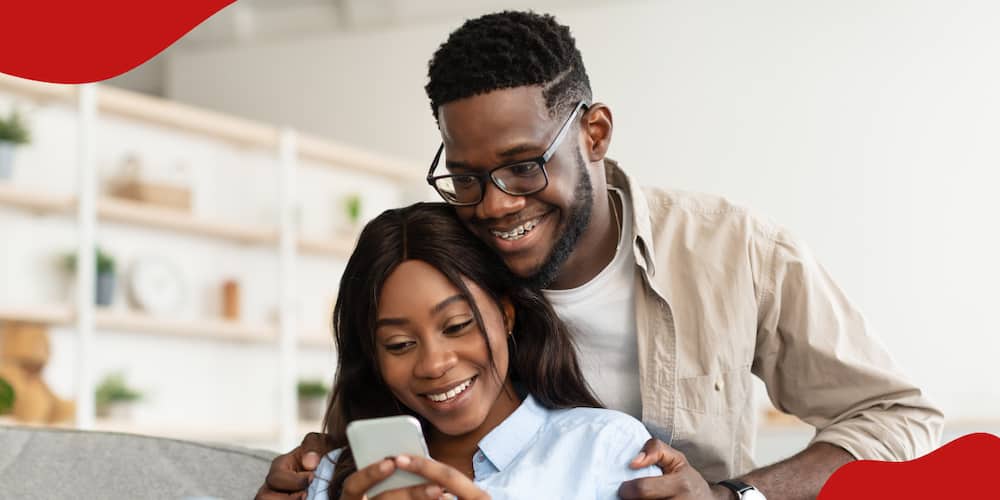Smiling young black man in braces embracing woman from behind while looking at smartphone.