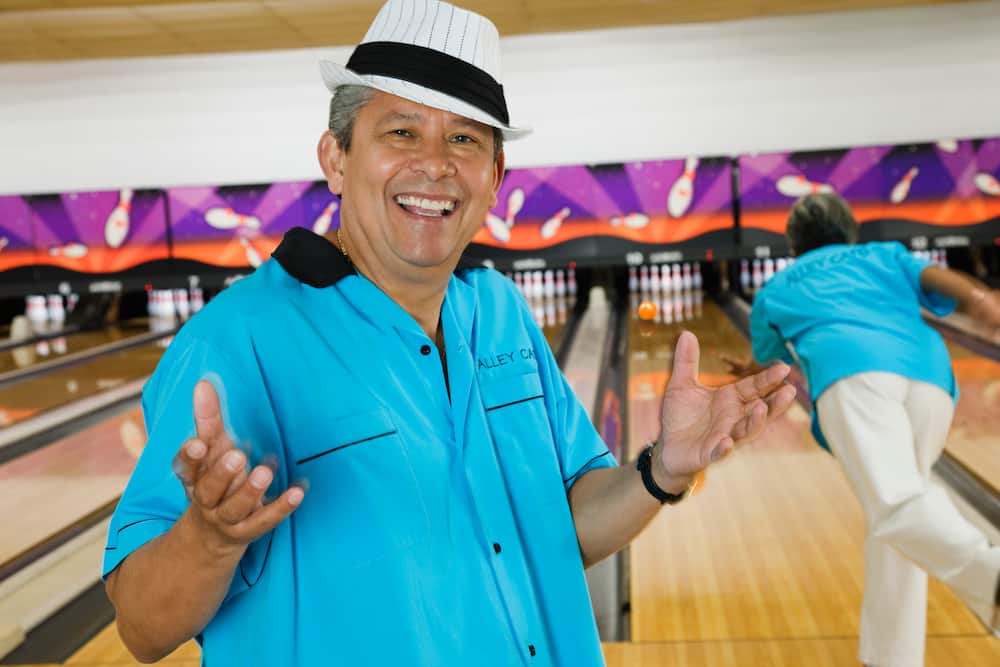 An elderly hispanic man in blue smiles at a bowling alley.