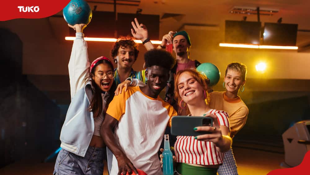 Close friends smile and take a group selfie while holding drinks and bowling balls.