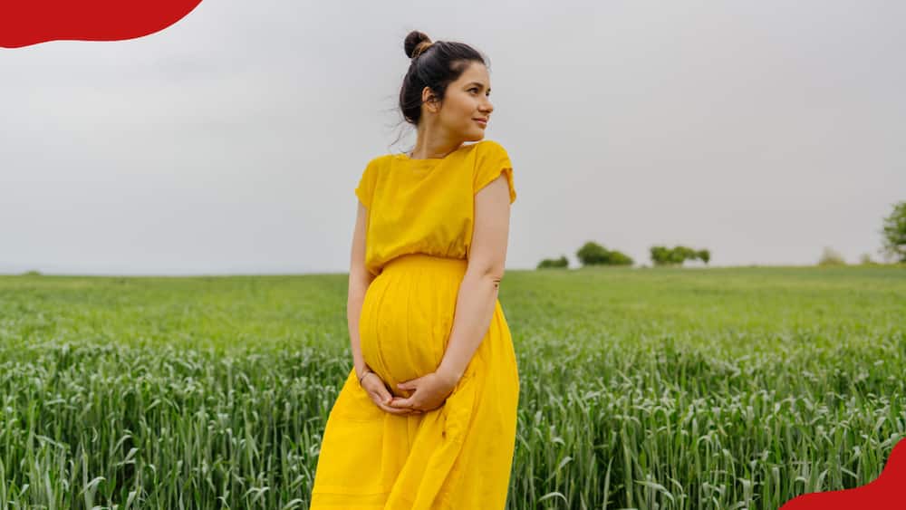 A pregnant woman is standing alone at the grass field