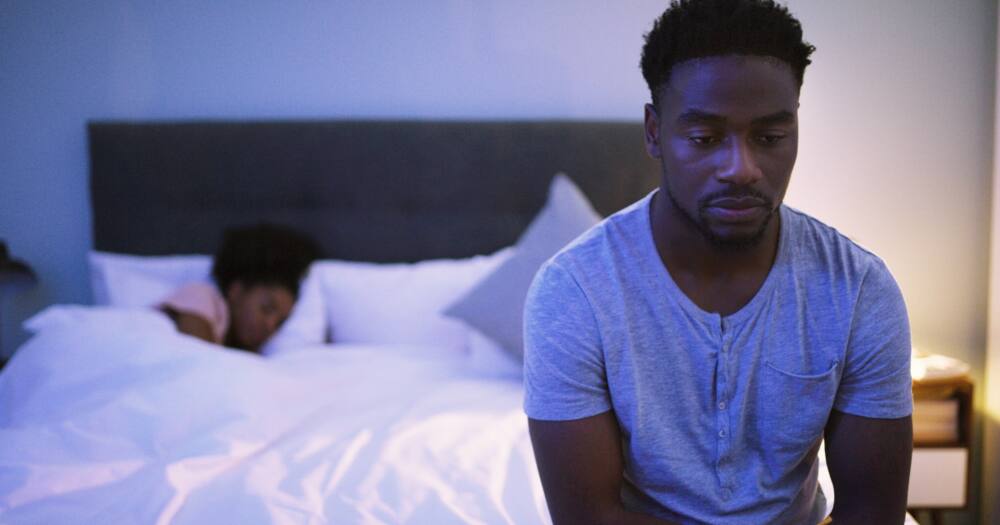 Shot of a concerned-looking young man sitting on a bed while his wife sleeps in the background. Photo: Getty Images.