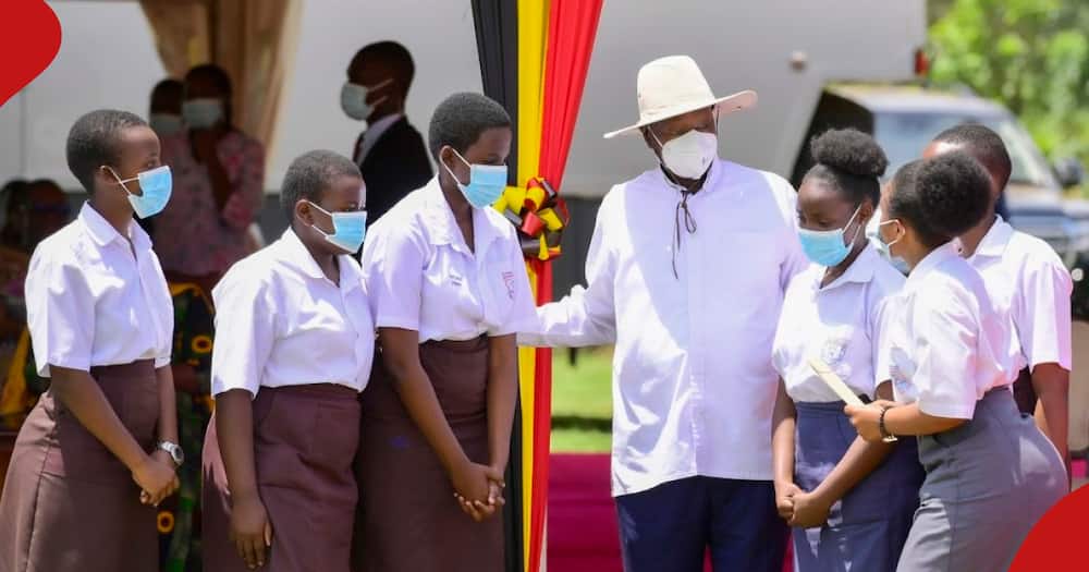 President Museveni, wearing a white hat, is surrounded by young students in uniform at an outdoor Labour Day celebration, marked by vibrant decorations and a spirit of community engagement.