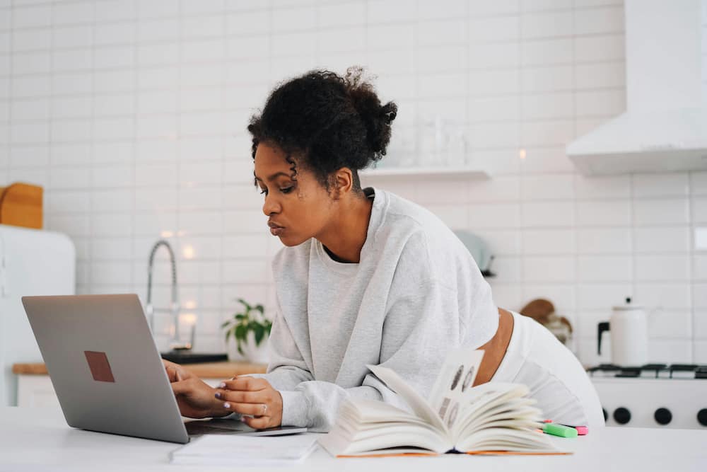 A woman in a white sweater is using a laptop at home, next to a book